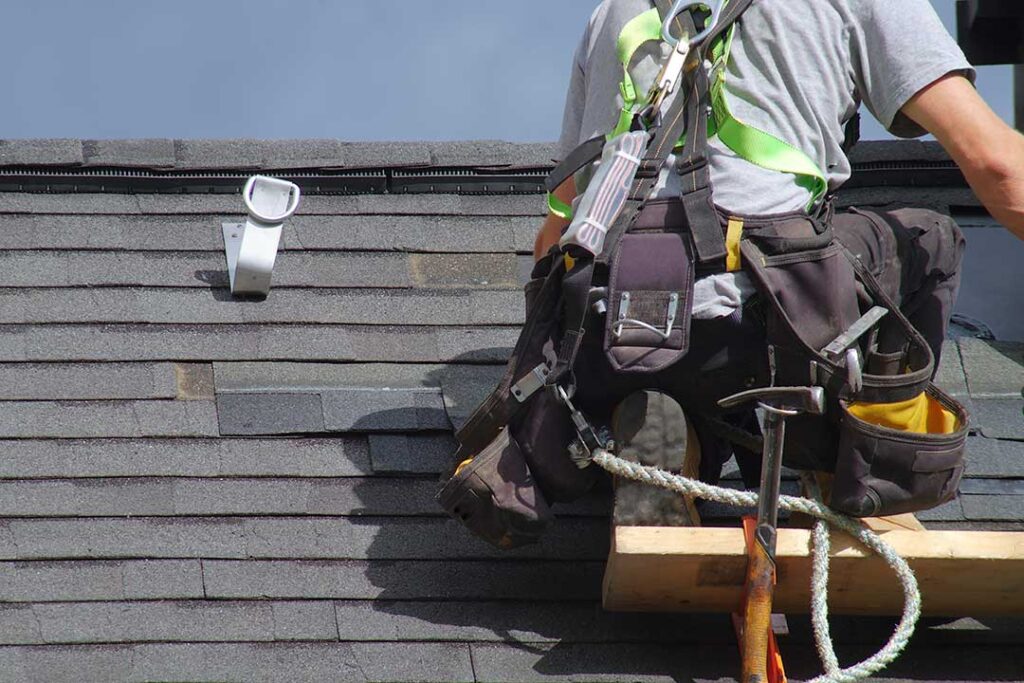 A homeowner stands in a yard using binoculars to inspect the condition of a roof surrounded by trees.