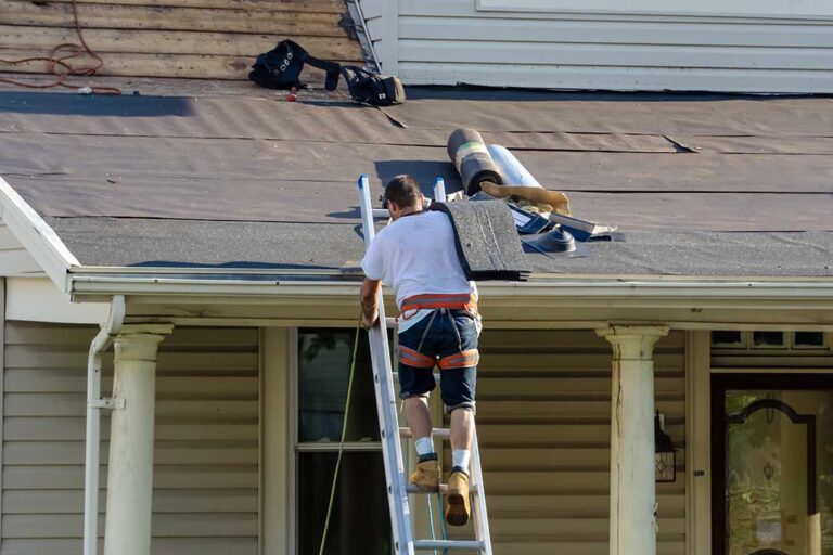 A homeowner examines aging asphalt shingles and roof flashing beside a snow-dusted residential house.