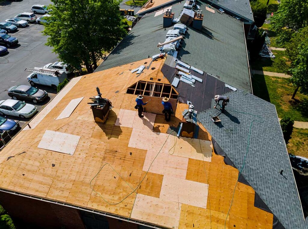 A homeowner reviews a roofing contract beside roof damage photographs and a calculator at a kitchen table.