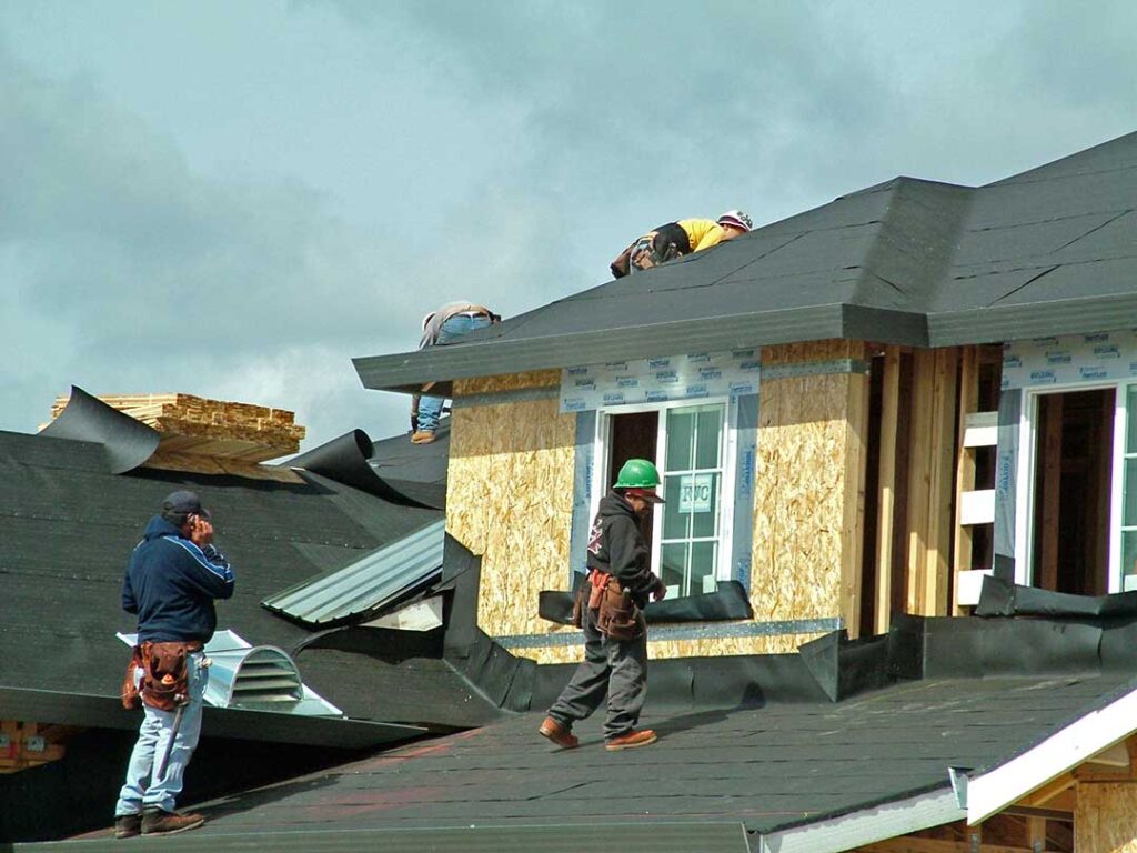 A homeowner examines curled shingles and roof flashing from the ground with binoculars beside a snow-dusted house.
