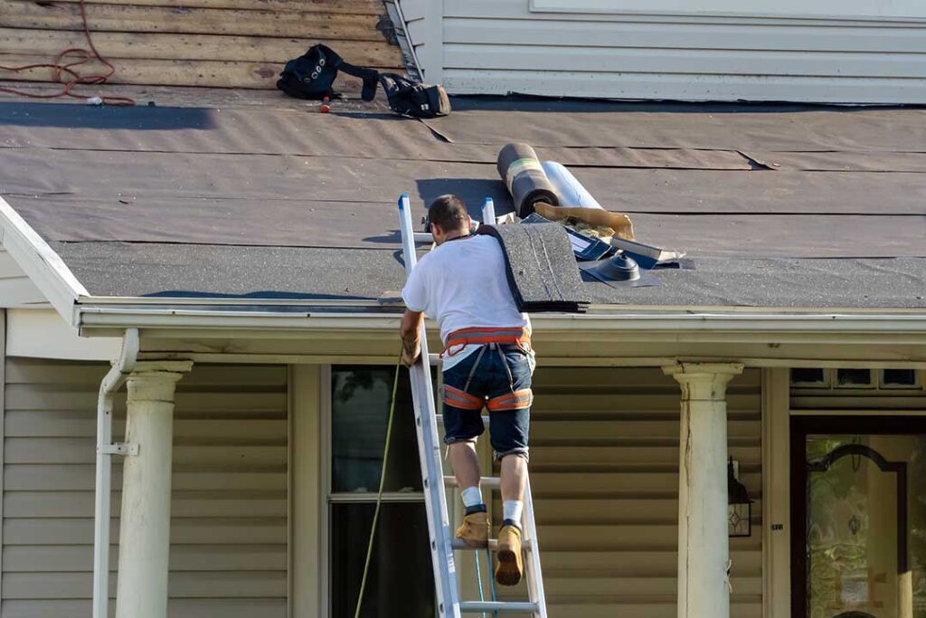 Roofing workers remove old shingles from a residential roof while protective tarps cover the landscaping below. KEYWORDS: ["roof tear-off workers", "old shingles removal", "residential roof deck"]
